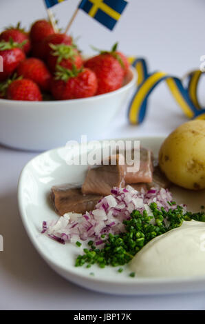 Eingelegter Hering mit neuen Kartoffeln, Schnittlauch, roten Zwiebeln und Sauerrahm auf einem Porzellanteller. Eine Schale mit Erdbeeren auf der Seite. Stockfoto