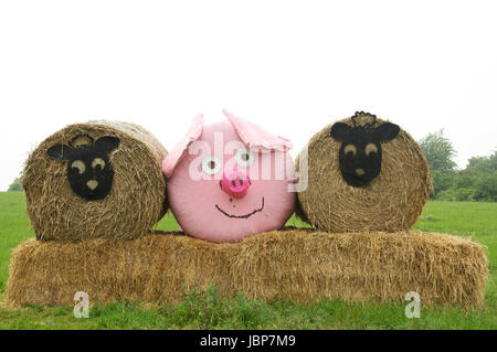 Comic-Tiergesichtern Vertretung Schafe und ein rosa Schwein schmücken drei Heuballen, angeordnet in einem Feld in der Nähe von Dorchester. Dorset, England, Vereinigtes Königreich. Stockfoto