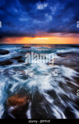 Dunkle Dämmerung Zeitpunkt Bungan Strand der Pazifikküste in Sydney, Australien. Starke Wellen Rollen auf erodierten Felsen unter orange der aufgehenden Sonne. Stockfoto