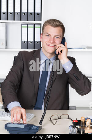 Junger Geschäftsmann Sitzt bin Schreibtisch Im Büro Und Wählt Eine Nummer bin Telefon. Im Hintergrund Befindet Sich Ein Regal. Der Mann Schaut Zur Kamera Und Lächelt. Stockfoto