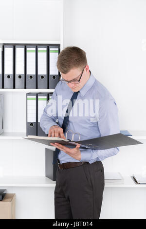 Junger Geschäftsmann Mit Brille Steht Vor Einem Regal Und Studiert Konzentriert Eine Akte. Der Mann Schaut Zur Akte. Stockfoto