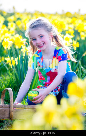 Glückliches kleine Mädchen ist für die Kamera Lächeln, während einigen Eiern in ihrem Körbchen zu sammeln, während eine Ostereiersuche. Sie ist in einem Feld voller Narzissen. Stockfoto