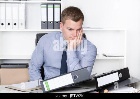 Junger Nachdenklicher Geschäftsmann Sitzt Vor Einem Berg von Akten Auf seit Schreibtisch Im Büro. Im Hintergrund Befindet Sich Ein Regal. Stockfoto