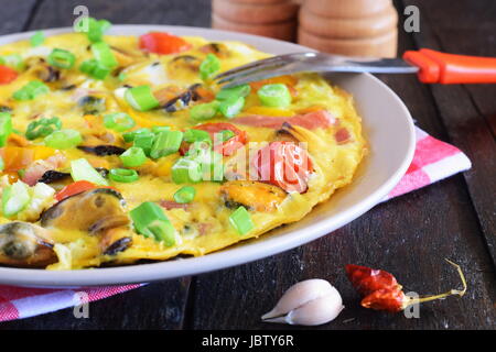 Omelett mit Muscheln, Cherry-Tomaten und Frühlingszwiebeln auf einer keramischen Platte auf einem schwarzen Hintergrund. Stockfoto