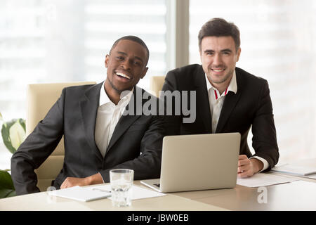 Zwei fröhliche Geschäftsleute in Anzügen sitzen am Schreibtisch, Blick in die Kamera, freundliche Unternehmer bereit für Zusammenarbeit, erfolgreiches Unternehmen Stockfoto