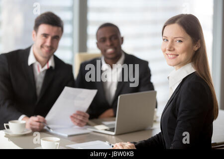 Attraktive junge Geschäftsfrau Lächeln für die Kamera sitzt am Schreibtisch, junge Führungskraft hält Geschäftstreffen für schwarze und weiße Untergebenen c Stockfoto