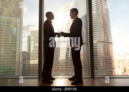 Zwei lächelnde schwarz-weiß Jungunternehmer Handshaking Stand in der Nähe großer Fenster mit Stadtgebäude außerhalb zuversichtlich kaukasischen und Afro-Amerikaner Stockfoto