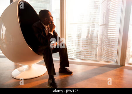 Seitenansicht des afrikanischen amerikanischen Geschäftsmann hält Hand am Kinn sitzen in Ei Designstuhl Sonnenuntergang, Blick durch große Bürofenster im city Stockfoto