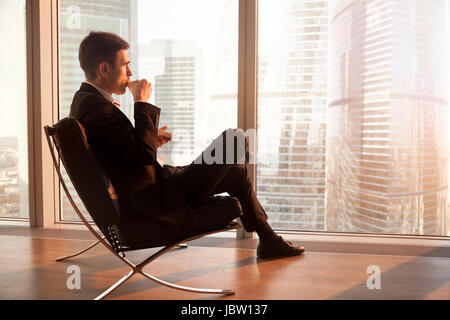 Seitenansicht des gut aussehend Geschäftsmann sitzen in bequemen Büro Stuhl genießen Tasse Kaffee am Morgen, Blick durch große Fenster im Morgengrauen Sonnenaufgang Stockfoto