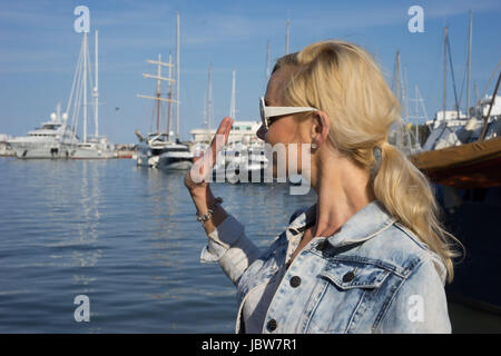 Attraktive blonde Frau mit Sonnenbrille mit ihrem Haar in einem Pferdeschwanz stehen winken mit Blick auf einen Hafen voller Luxus Sportboote und Yachten Stockfoto