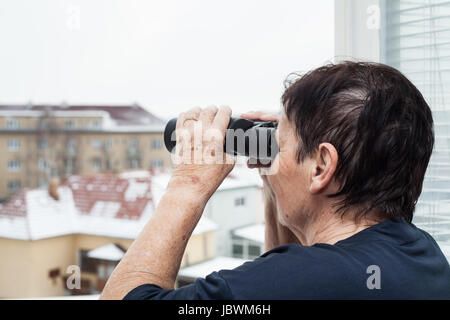 Ältere Frau mit dem Fernglas mit Blick vom Fenster. Stockfoto