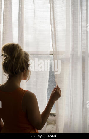 Rückansicht einer blonden kaukasischen Frau steht am Fenster, wegschauen Stockfoto