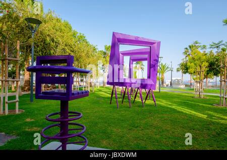 Ein Blick auf Molos Promenade an der Küste von Limassol Stadt in Zypern. Ein Blick auf eine moderne lila Kunst-Skulptur des quadratischen Rahmen und einen Stuhl, der zu Fuß Weg, umgeben von Palmen, Rasen und das Mittelmeer. Stockfoto