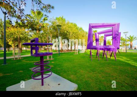 Ein Blick auf Molos Promenade an der Küste von Limassol Stadt in Zypern. Ein Blick auf eine moderne lila Kunst-Skulptur des quadratischen Rahmen und einen Stuhl, der zu Fuß Weg, umgeben von Palmen, Rasen und das Mittelmeer. Stockfoto