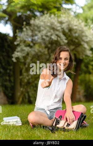 Fröhliche Studentin auf dem Rasen sitzen zeigt Daumen oben Sommer Stockfoto