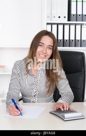 Eine Junge, Lächelnde Frau Führt Mit Einem Tischrechner Berechnungen Durch Und Macht Sich so Mit Einem Blauem Stift Auf Einem Blatt Papier bin Schreibtisch Im Büro. Im Hintergrund Steht Ein Regal. Sterben Sie Frau Schaut Zur Kamera. Stockfoto