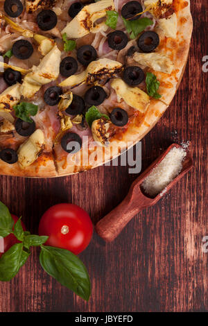 Rustikal, Country Style Pizza Hintergrund. Pizza mit schwarzen Oliven, Schinken und Artischocken auf hölzernen Hintergrund mit Parmesan-Käse, Tomaten und Basilikum. Stockfoto