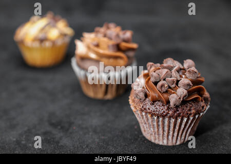 Drei Schokoladen Cupcakes garniert mit Schokolade-Chips und Schokolade Zuckerguss auf aufgereiht über den Rahmen auf schwarzem Hintergrund, von rechts nach links Stockfoto