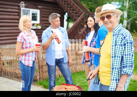 Porträt von senior glücklich Würstchen braten und auf dem Lande am Wochenende mit Gruppe junger Freunde sprechen auf Hintergrund betrachten Stockfoto