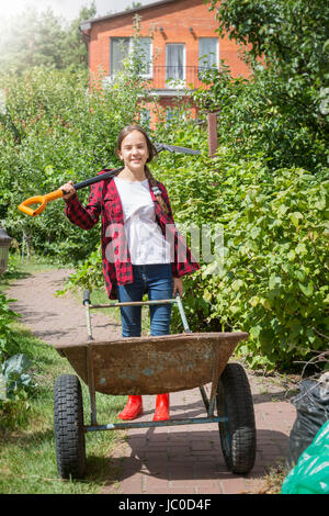 Porträt von fröhliches junges Mädchen posiert mit Schubkarre und Schaufel in Garten am sonnigen Tag Stockfoto