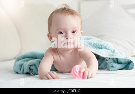 Porträt von entzückenden Baby unter blaues Handtuch auf dem Bett liegen und halten Plastikspielzeug Stockfoto