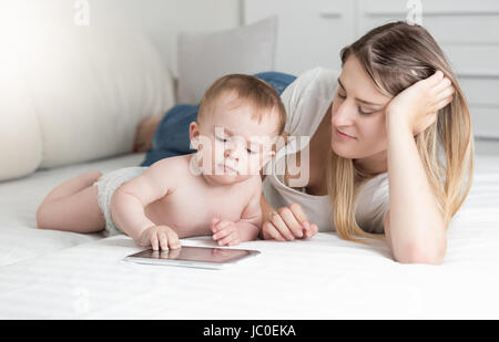 Porträt der schönen lächelnden Mutter und 10 Monate alten Baby jungen auf Bett liegend mit Tablet-PC Stockfoto