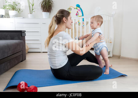 Schöne lächelnde Mutter praktizieren Yoga auf Fitness-Matte im Wohnzimmer Stockfoto