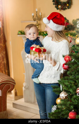 Porträt der glückliche junge Mutter im Wohnzimmer neben Weihnachtsbaum anwesend sein, um ihr Baby Sohn auf Weihnachten geben Stockfoto