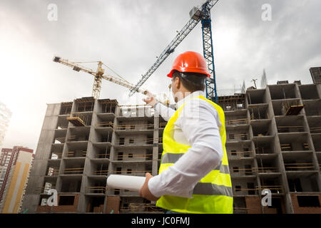 Porträt des jungen Architekten halten Blaupausen und deutete auf Gebäude im Bau Stockfoto