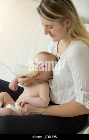 Porträt der jungen Mutter auf ihr Baby Flasche verleiht Bett sitzend getönt Stockfoto