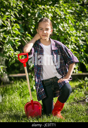 Niedliche Mädchen in Gummistiefeln posiert mit Spielzeug Spaten im Garten Stockfoto