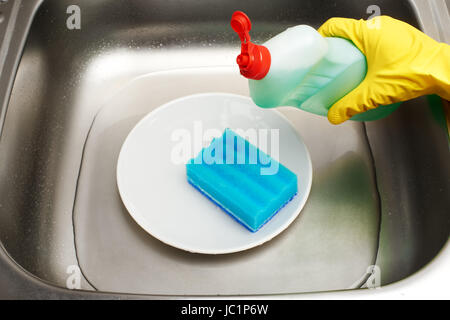 Nahaufnahme der Küchenspüle mit Wasser, saubere weiße Platte, blaue Reinigungs Schwamm und Flasche Spülmittel in weiblicher Hand in gelben Gummischutz Stockfoto