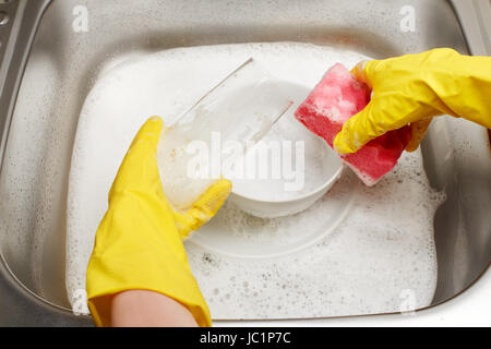 Nahaufnahme von weiblichen Händen schützende Gummihandschuhe Trinkglas mit rosa Reinigung Waschen gegen Küchenspüle voller Schaum und Registerkarte "sponge gelb Stockfoto