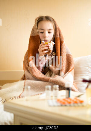 Porträt des kleinen Mädchens bedeckt im Karomuster am Bett sitzen und Tee trinken Stockfoto