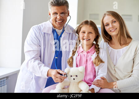 Lächelnde Mädchen mit Teddybär, ihre Mutter und Arzt Blick in die Kamera in Kliniken Stockfoto