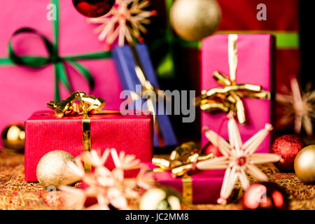 Crimson Geschenkbox mit goldenen Bogen zwischen Kugeln und Stroh Sterne. Fünf weitere einfarbig Pakete außerhalb des Fokus. Hintergrund für Weihnachten Komposition. Stockfoto