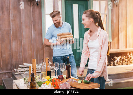 Lächelnde junge Frau schneiden Gurke und bärtigen Mann mit Brennholz für bbq Stockfoto