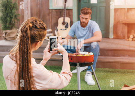 Frau nehmen Foto des Mannes Grillen von Fleisch auf Veranda Stockfoto