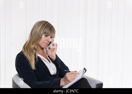 Eine Frau Telefoniert Mit Einem Smartphone Und Macht Dabei Notizen Auf Einem Blatt Papier, füttere Sie in Einem Sessel Sitzt. Sterben Sie Frau Schaut Zur Seite. Stockfoto