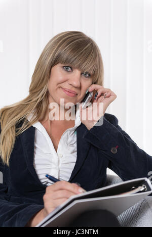 Eine Frau Telefoniert Mit Einem Smartphone Und Macht Dabei Notizen Auf Einem Blatt Papier, füttere Sie in Einem Sessel Sitzt. Sterben Sie Frau Schaut Zur Kamera. Stockfoto