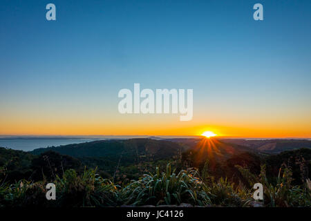 Sonnenuntergang über dem Hauraki-Golf, mit Bäumen und Hügeln Silhouette im Vordergrund. Aufgenommen am Strand von Whakanewha Camp Ground auf Waiheke ist Stockfoto