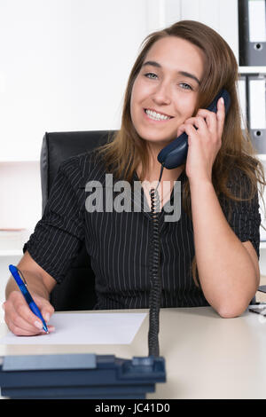 Eine Junge, Lächelnde Frau Telefoniert Und Macht Dabei Notizen Auf Einem Blatt Papier sind Sie bin Schreibtisch Im Büro Sitzt. Im Hintergrund Steht Ein Regal. Sterben Sie Frau Schaut Zur Kamera. Stockfoto