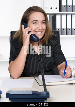 Eine Junge, Lächelnde Frau Telefoniert Und Macht Dabei so in Einem Aktenordner füttere Sie bin Schreibtisch Im Büro Sitzt. Im Hintergrund Steht Ein Regal. Sterben Sie Frau Schaut Zur Kamera. Stockfoto