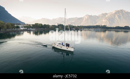 Luftbild auf einem Segelboot und Rescue Boot überqueren den Genfer See, mit Bergen im Hintergrund, Bäume am Ufer des Sees und eine Marina in Villeneuve, Kanton Waadt, Schweiz Stockfoto