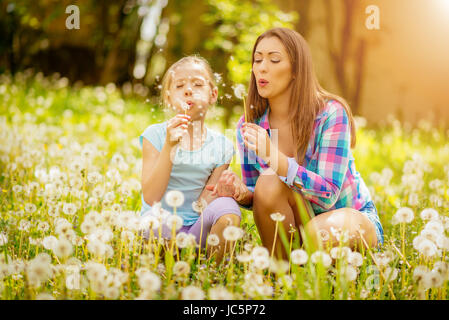 Glückliches kleines Mädchen bläst Löwenzahn mit Mutter im Park. Stockfoto