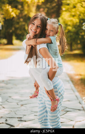 Schöne Mutter und Tochter ein Spaziergang durch einen Park im Frühling. Junge Mutter seiner Tochter Huckepack. Stockfoto