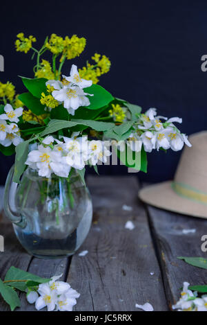 Stilllife-Karte mit Jasminblüten in Glas, separate Zweige mit Blüten, Blütenblätter und Strohhut auf dem rustikalen Holztisch. Weiche selektive focu Stockfoto
