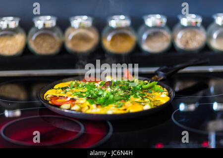 Omelette mit Gemüse auf elektrisches Herd und Schalen mit Gewürzen im Hintergrund der Küche. Stockfoto