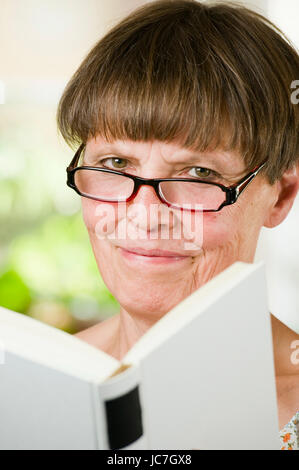 Innenaufnahme Kopf Und Schulter-Porträt Einer Älteren Frau Mit Lesebrille Und Offenem Buch Freundlich in Die Kamera lächelnd Stockfoto