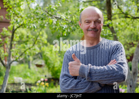 Reifer Mann sitzen im Garten in der Nähe von Apfelbaum im Lande. Er zeigt thump up. Stockfoto